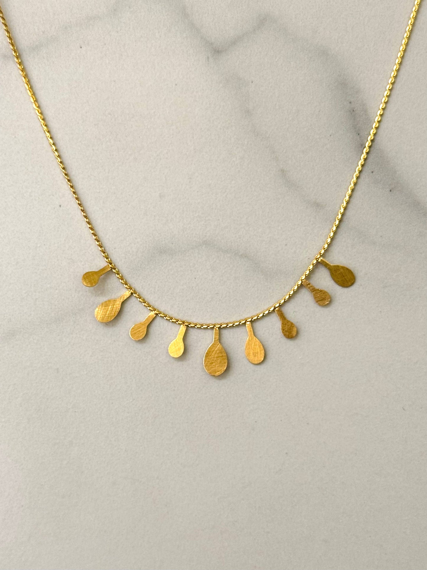 Collier ras-du-cou en acier inoxydable doré à chaîne fine orné sur le devant de neuf pampilles plates pendantes en forme de gouttes au fini texturé brossé, présenté sur un fond en marbre blanc.