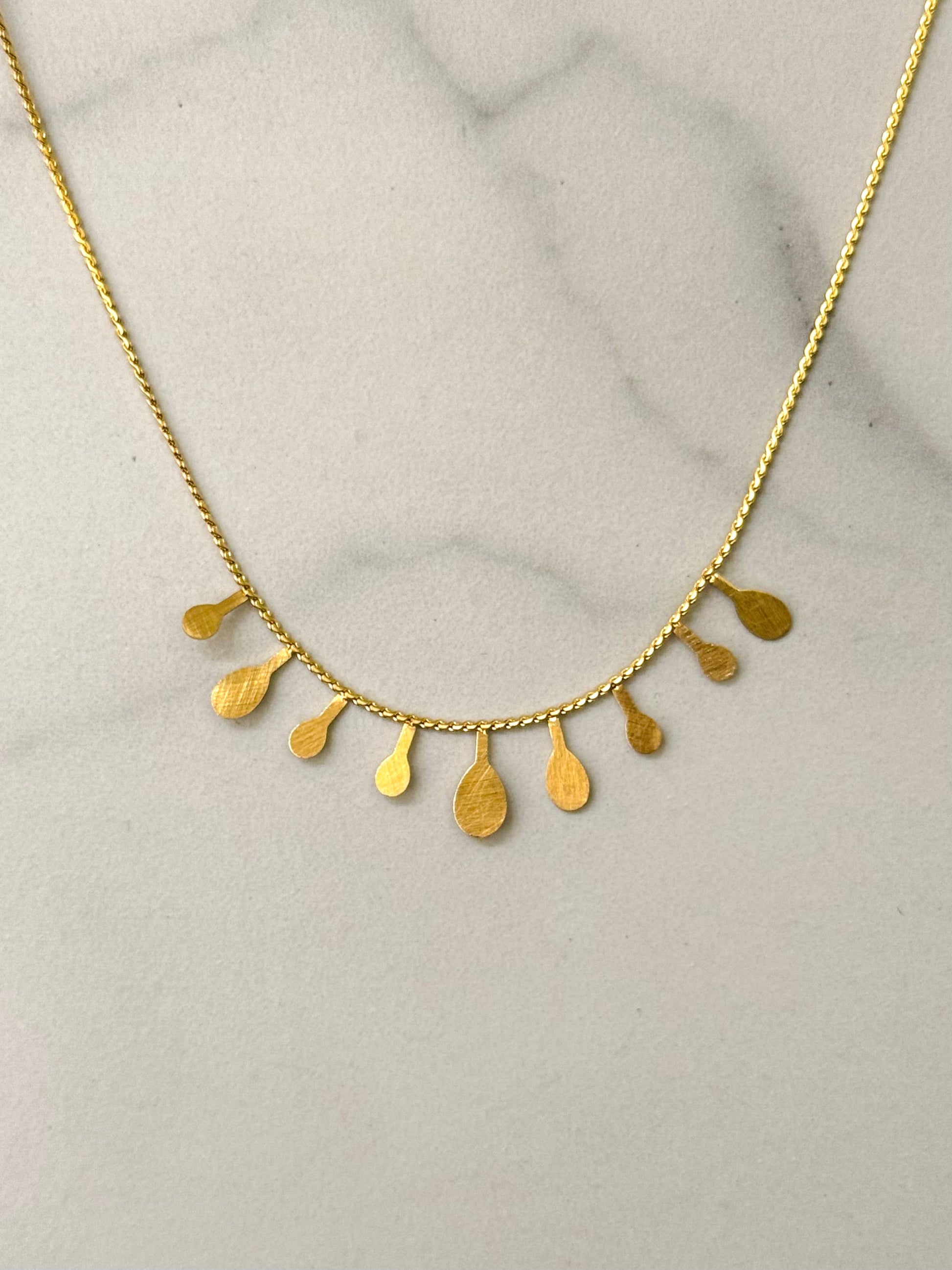 Collier ras-du-cou en acier inoxydable doré à chaîne fine orné sur le devant de neuf pampilles plates pendantes en forme de gouttes au fini texturé brossé, présenté sur un fond en marbre blanc.