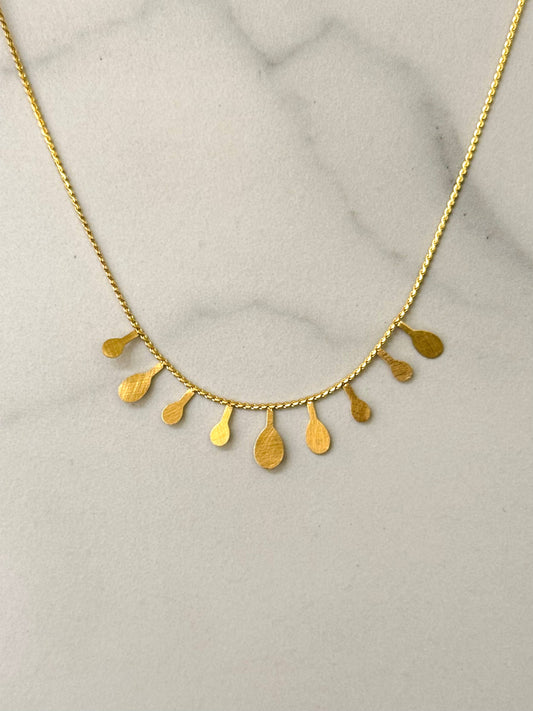 Collier ras-du-cou en acier inoxydable doré à chaîne fine orné sur le devant de neuf pampilles plates pendantes en forme de gouttes au fini texturé brossé, présenté sur un fond en marbre blanc.