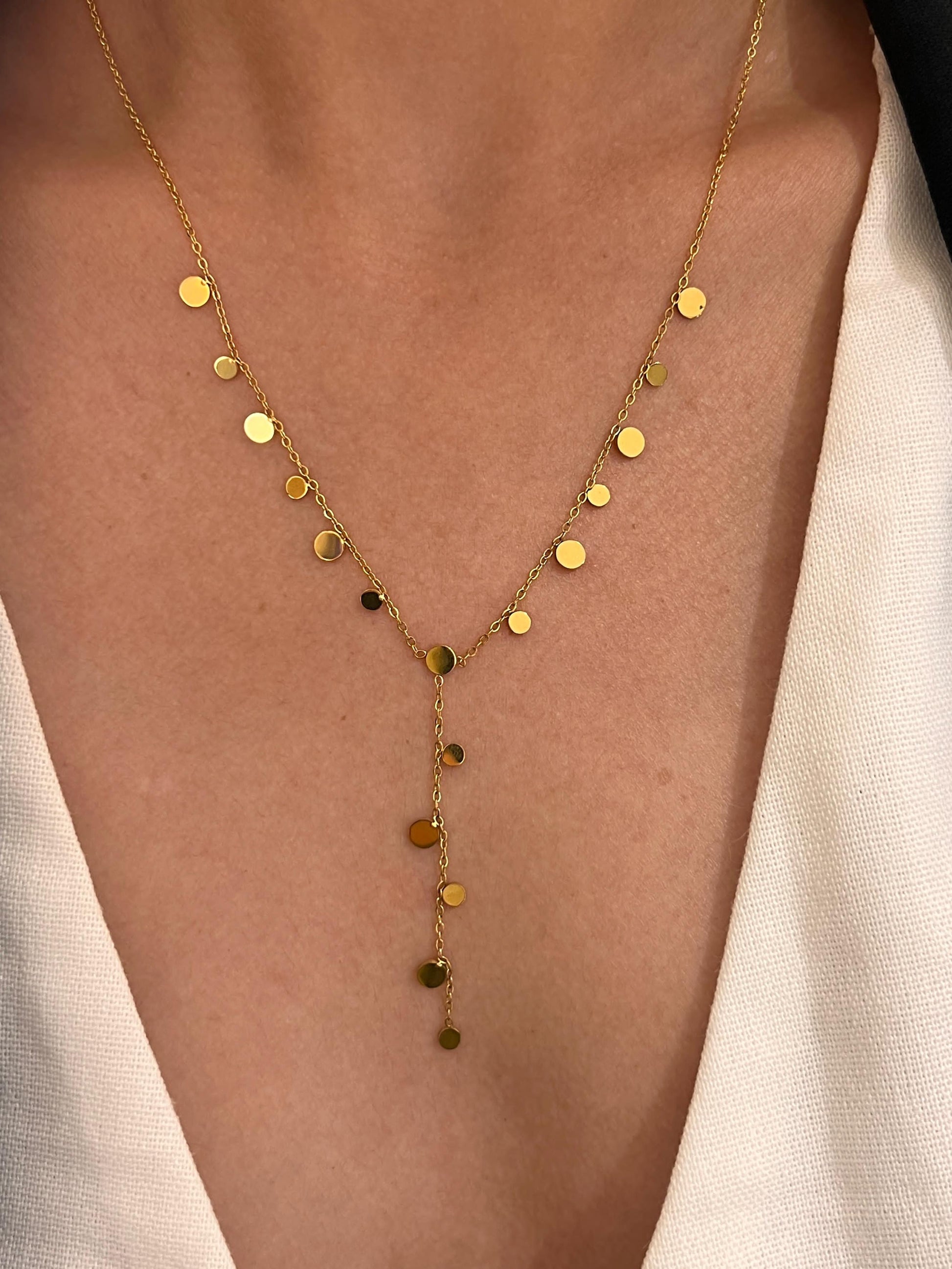 Collier style cravate en forme de Y en acier inoxydable doré, orné de nombreuses petites pastilles rondes et plates au fini miroir suspendues, porté sur le décolleté d'une femme habillée d'un vêtement blanc.