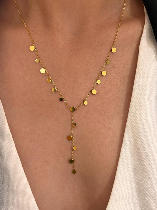 Collier style cravate en forme de Y en acier inoxydable doré, orné de nombreuses petites pastilles rondes et plates au fini miroir suspendues, porté sur le décolleté d'une femme habillée d'un vêtement blanc.