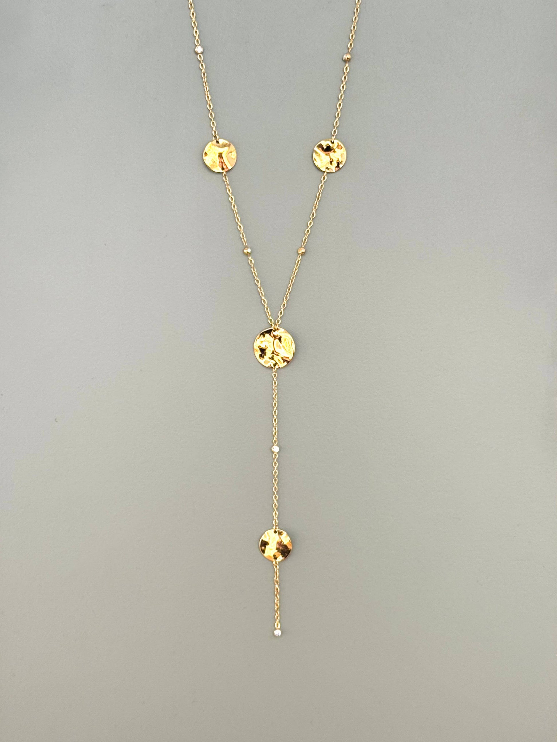 Collier style cravate en forme de Y en acier inoxydable doré, orné de quatre pastilles rondes ciselées (texturées) et de perles de diamant, suspendues verticalement sur un fond gris neutre.