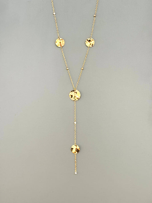 Collier style cravate en forme de Y en acier inoxydable doré, orné de quatre pastilles rondes ciselées (texturées) et de perles de diamant, suspendues verticalement sur un fond gris neutre.