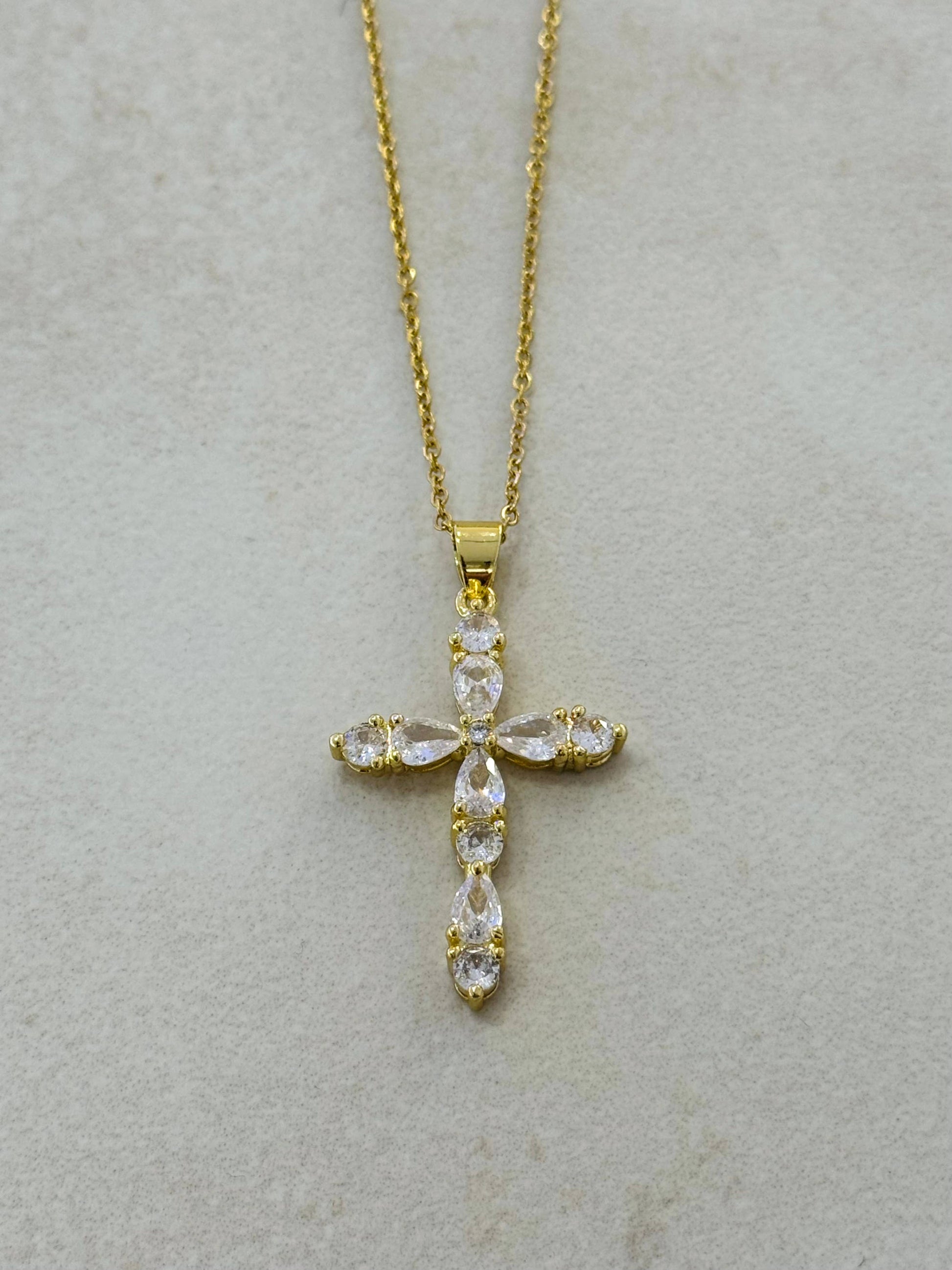 Collier en acier inoxydable doré avec une fine chaîne et un grand pendentif en forme de croix baroque pavée de cristaux d'oxydes de zirconium taillés en poire et ronds, posé à plat sur un fond beige texturé.