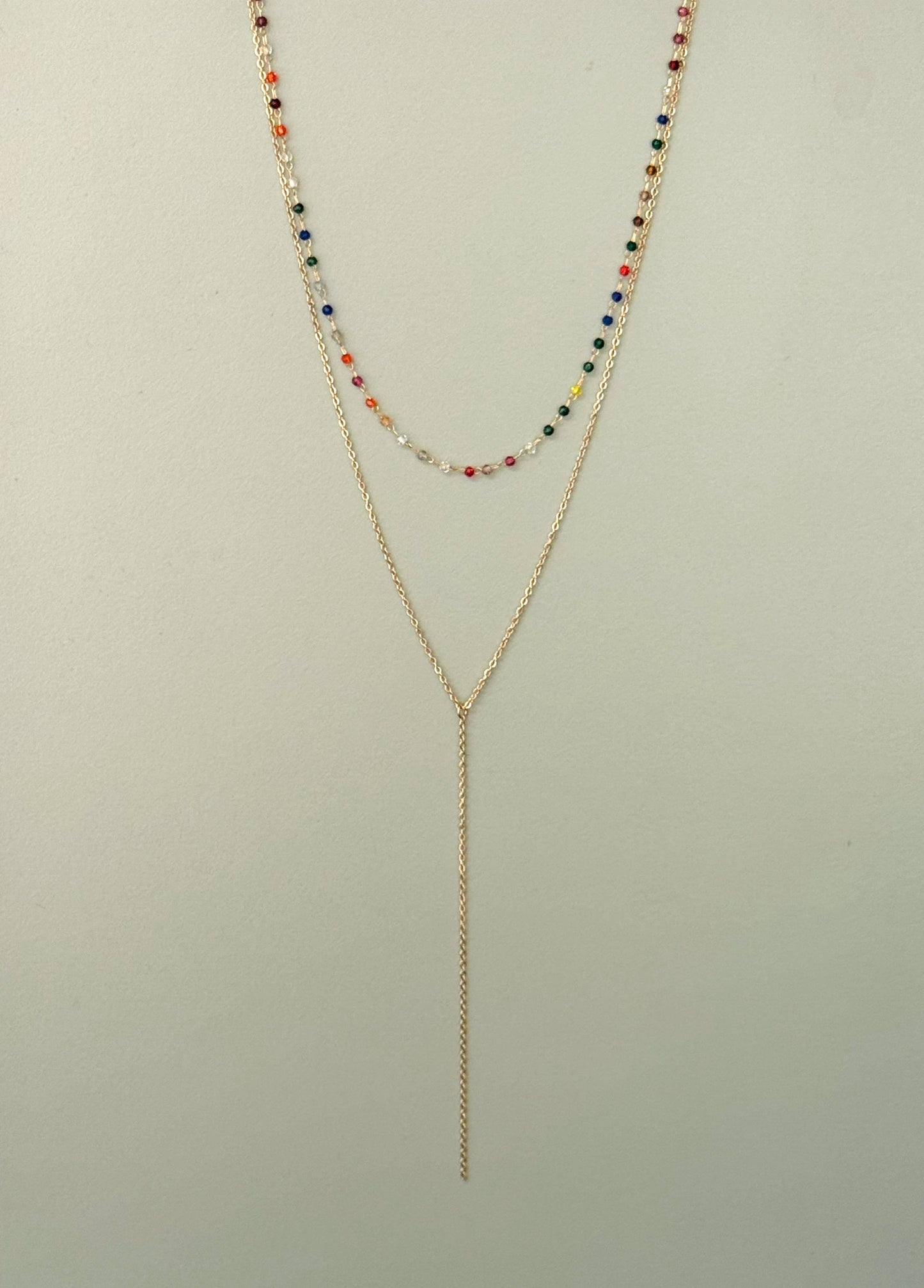 Collier double rang en acier inoxydable doré composé d'un premier rang ras-du-cou avec de petites perles multicolores et d'un second rang en chaîne fine plongeante en forme de Y style cravate, présenté de face sur fond vert de gris clair.