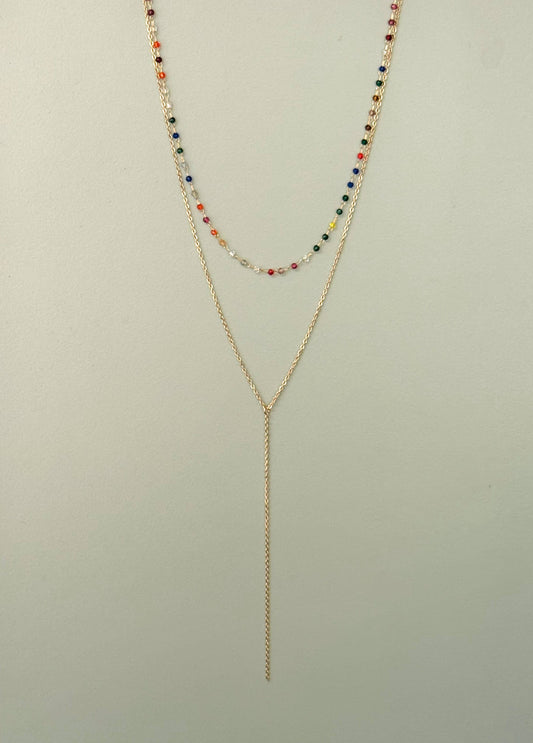 Collier double rang en acier inoxydable doré composé d'un premier rang ras-du-cou avec de petites perles multicolores et d'un second rang en chaîne fine plongeante en forme de Y style cravate, présenté de face sur fond vert de gris clair.