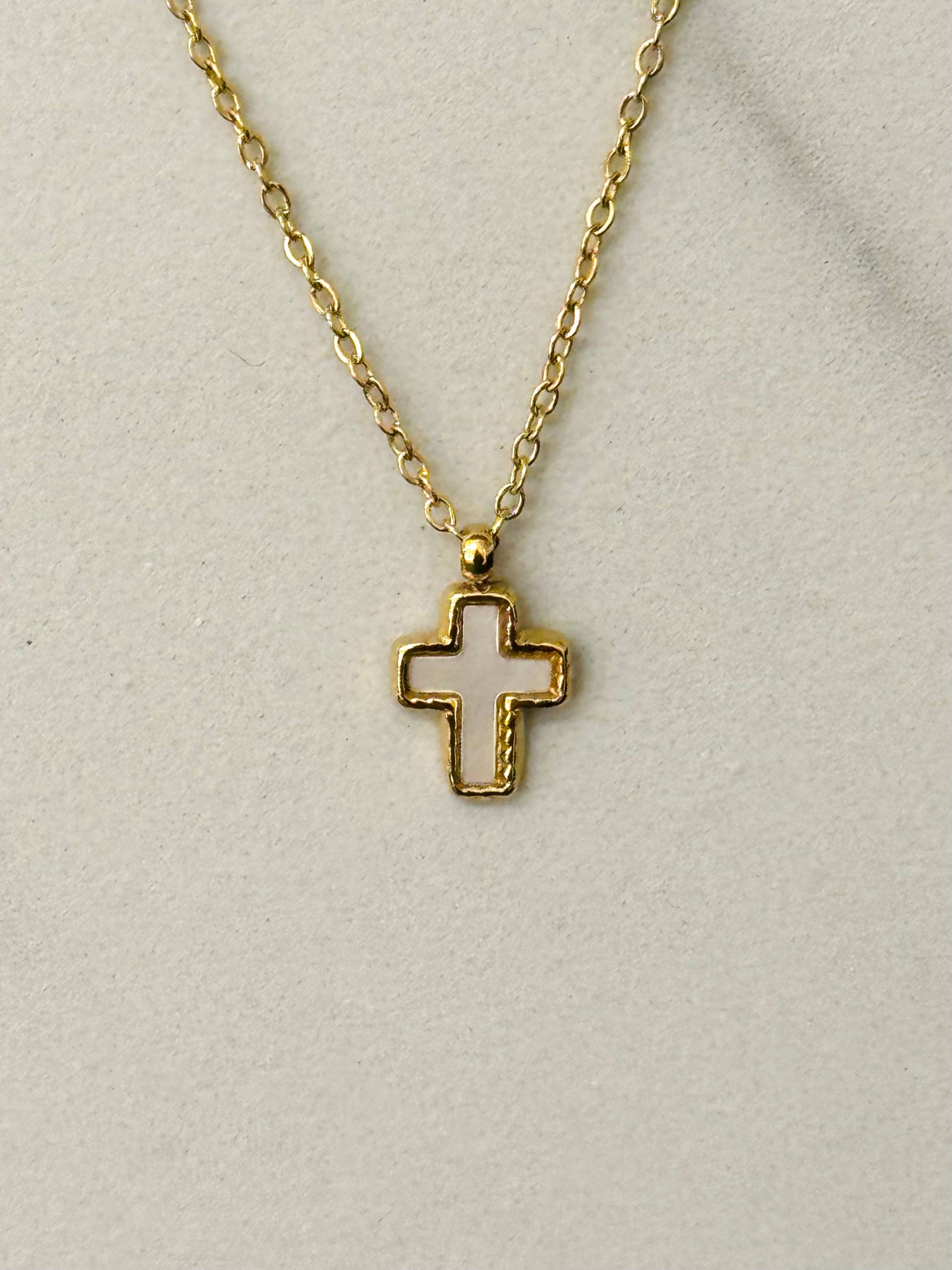Gros plan sur un collier en acier inoxydable doré avec une fine chaîne et un pendentif petite croix orné d'un cœur en nacre blanche irisée, posé à plat.
