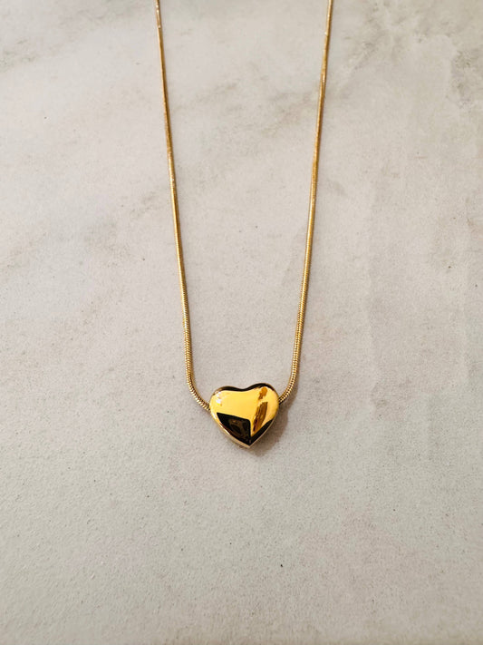 Collier en acier inoxydable doré composé d'une fine chaîne en maille serpent souple et d'un pendentif en forme de cœur bombé au fini lisse et poli miroir, posé à plat sur un fond texturé beige clair.