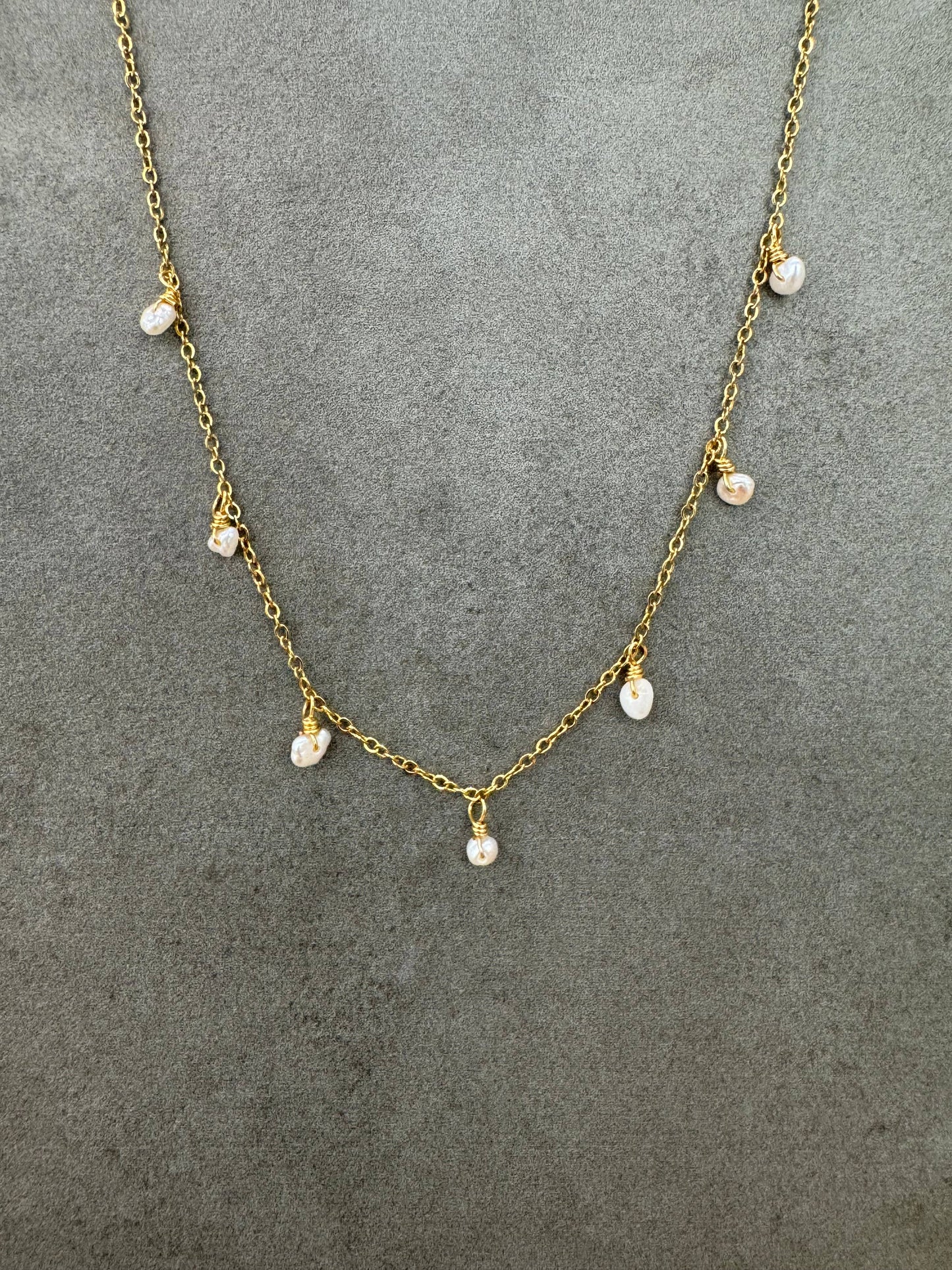 Collier fin en acier inoxydable doré à maille forçat, orné sur le devant de sept petites perles d'eau douce naturelles blanches et baroques suspendues en pampilles, présenté de face sur un fond gris texturé.