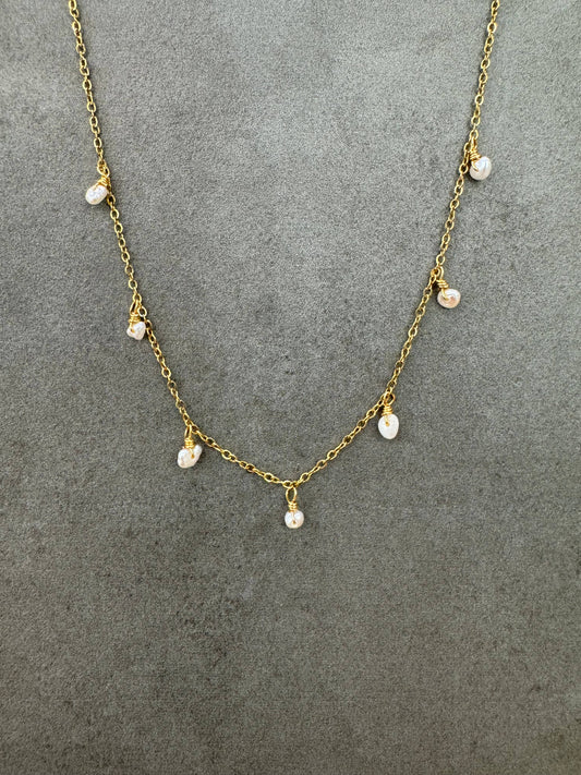 Collier fin en acier inoxydable doré à maille forçat, orné sur le devant de sept petites perles d'eau douce naturelles blanches et baroques suspendues en pampilles, présenté de face sur un fond gris texturé.