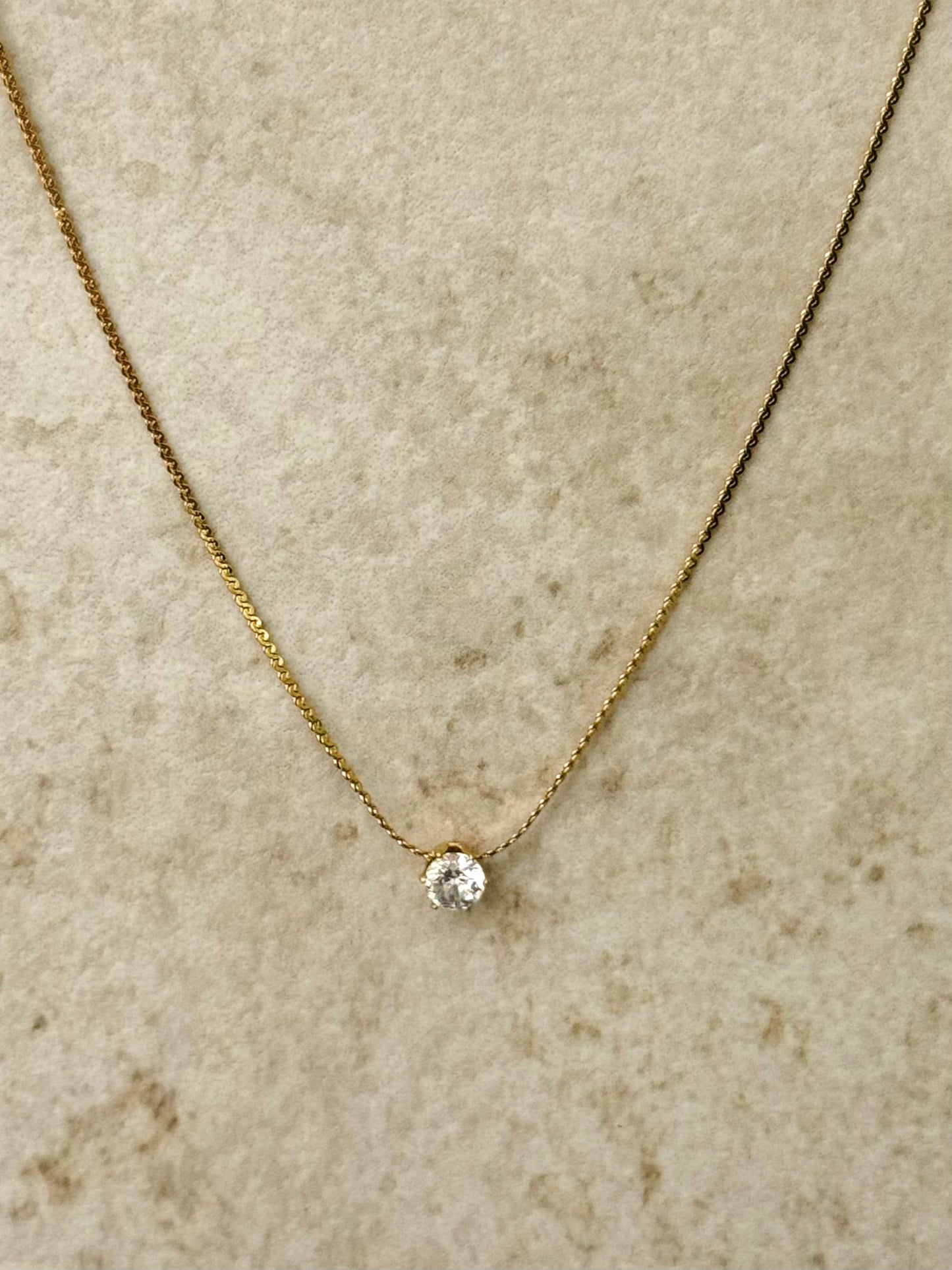 Collier à fine chaîne dorée en acier inoxydable orné d'un unique pendentif en cristal rond brillant de type solitaire, centré de face sur un fond beige texturé.