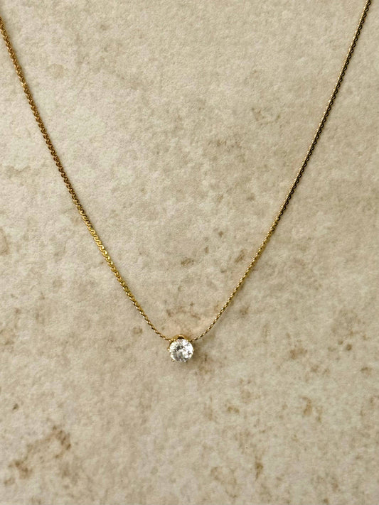 Collier à fine chaîne dorée en acier inoxydable orné d'un unique pendentif en cristal rond brillant de type solitaire, centré de face sur un fond beige texturé.