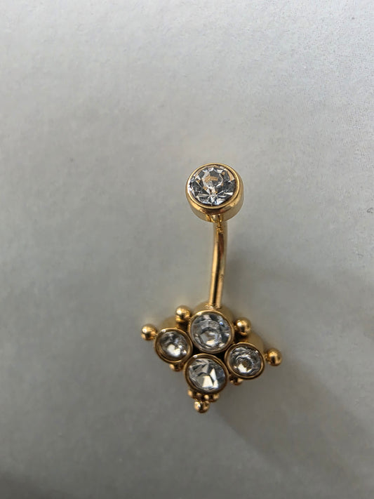 Piercing de nombril en acier inoxydable doré avec une tige courbée, une bille supérieure incrustée d'un cristal blanc et un motif inférieur fixe en forme de losange orné de quatre oxydes de zirconium ronds et de petites billes dorées, sur un fond gris épuré.